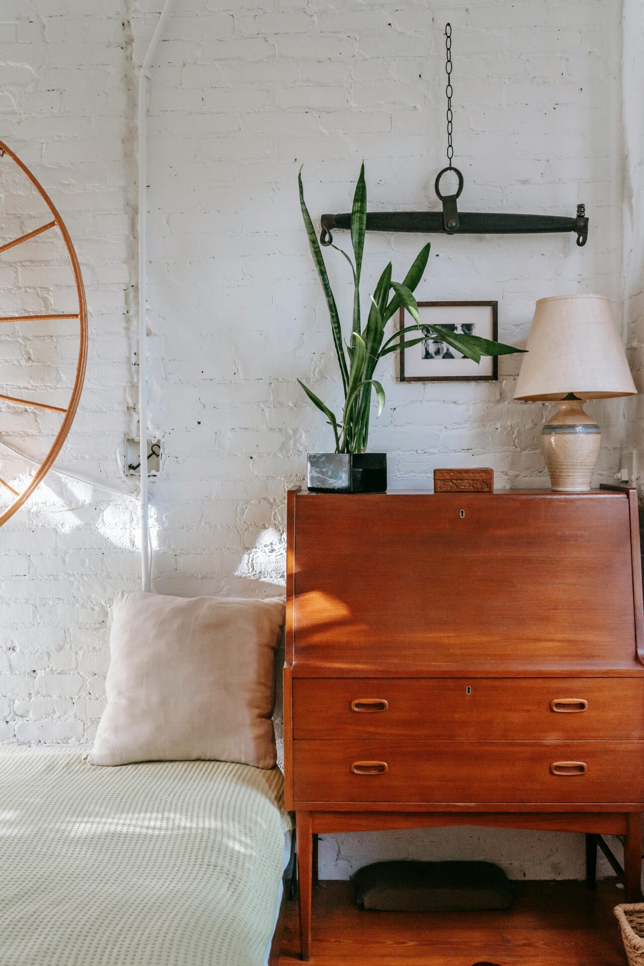 Light bedroom interior furnished with soft bed and vintage wooden cabinet decorated with lush green plant and lamp